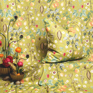 A close-up of a patterned fabric product, likely a bedspread or sofa cover, with a matching decorative pillow. The fabric has a light olive-yellow base with an all-over botanical print featuring white flowers, green leaves, peach-colored fruits, and small red berries.